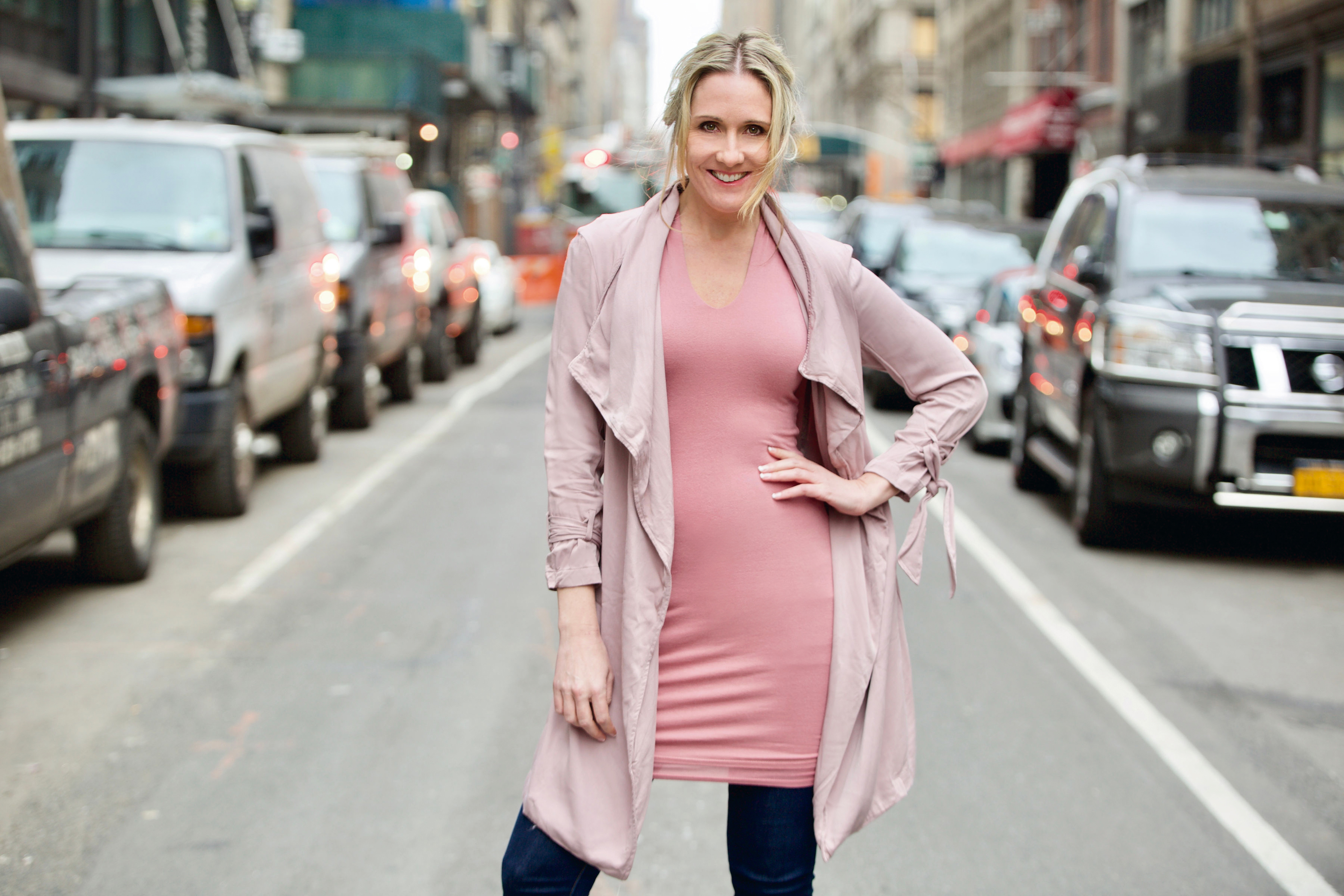 A woman in pink dress and jacket standing on the side of street.